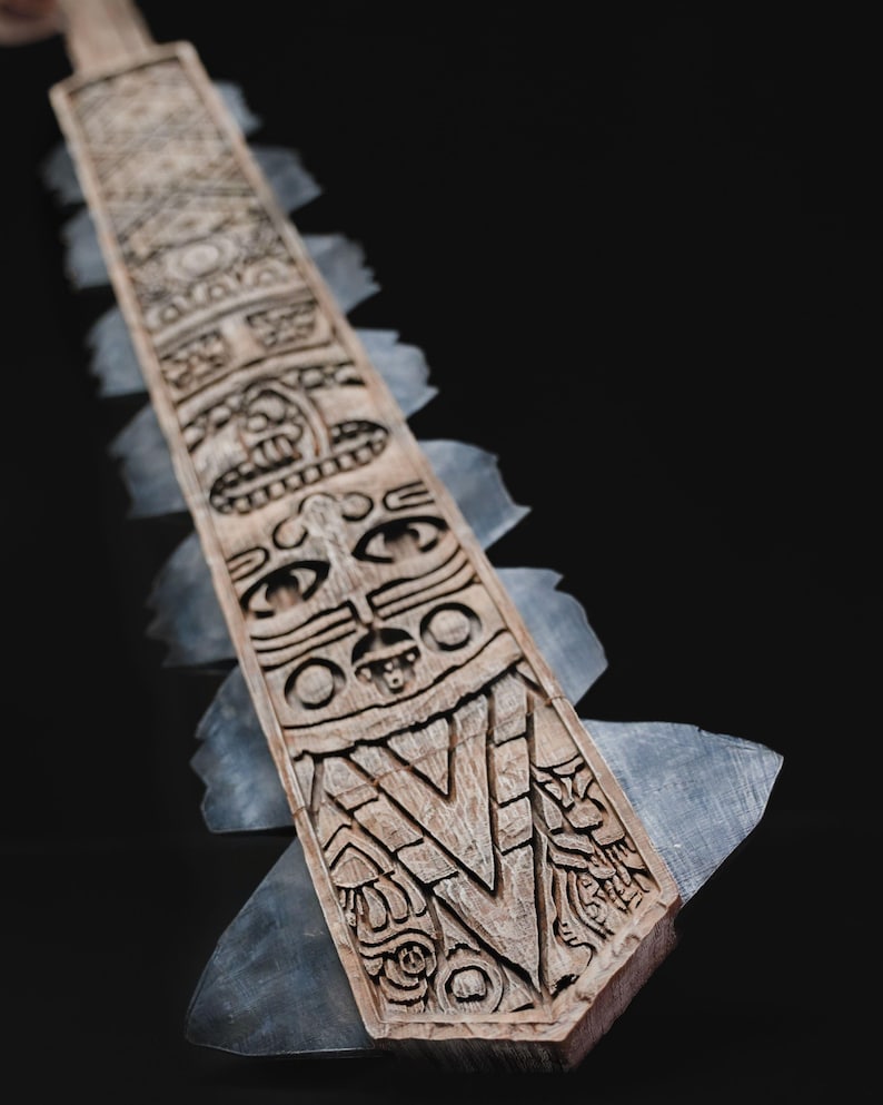 May include: A carved wooden paddle with intricate designs and a series of stylized faces. The paddle has a light brown color and is edged with translucent, gray-blue blades. The artwork is likely inspired by indigenous art.