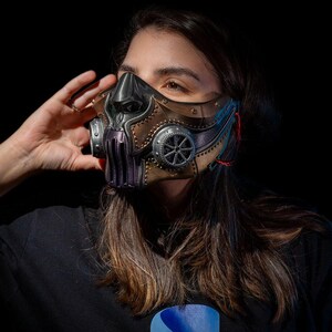May include: A steampunk-style mask with a metallic finish. The mask features a combination of bronze, silver, and purple elements, with intricate gear-like details and a sculpted face design. The mask is designed to cover the nose and mouth.