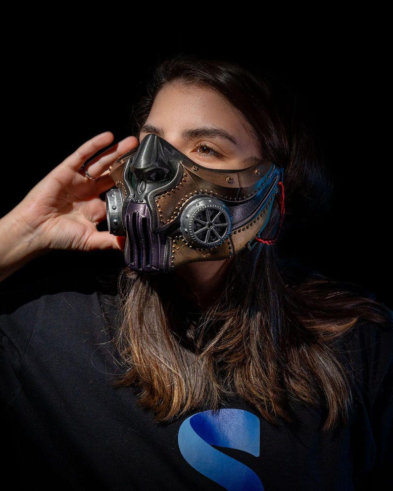 May include: A steampunk-style mask with a metallic finish. The mask features a combination of bronze, silver, and purple elements, with intricate gear-like details and a sculpted face design. The mask is designed to cover the nose and mouth.