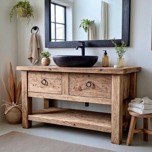 May include: A rustic wooden bathroom vanity with a black vessel sink, two drawers, and a lower shelf. The vanity has a natural wood finish and is paired with a black framed mirror, a small wooden stool, and a woven basket. The room is decorated with plants.