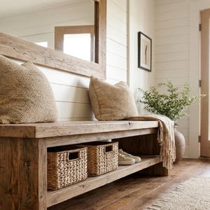 May include: A rustic wooden bench featuring a lower shelf with two woven baskets and a pair of sneakers. Two textured beige pillows rest on the seat, accompanied by a draped throw blanket. The bench is positioned below a large wooden-framed mirror.
