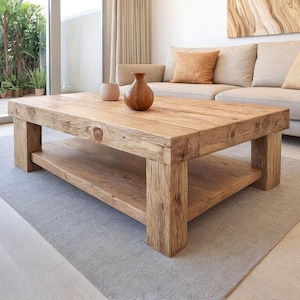 Peut inclure: Une table basse rectangulaire en bois massif avec une étagère inférieure. La table est en bois clair et a un aspect naturel non fini. Deux vases décoratifs sont posés dessus. La table est posée sur un tapis gris dans un salon.