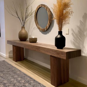 May include: A wooden console table with two legs on each side. The table has a natural wood finish and is decorated with a vase of dried branches, a vase of dried pampas grass, and a round mirror with a wooden frame.