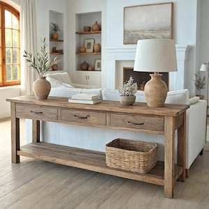 Peut inclure: Une console en bois rustique avec trois tiroirs et une étagère inférieure. Un panier en osier est posé sur l'étagère. La table est surmontée d'une lampe, d'un vase et d'objets décoratifs. La pièce a une palette de couleurs neutres.