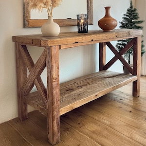 May include: A rustic wooden console table with a distressed finish. The table has a single shelf and a decorative X-shaped support structure. A vase with dried pampas grass, a candle holder, and a ceramic vase are displayed on the table.