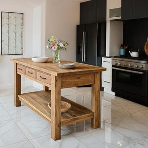 May include: A solid wood kitchen island with a natural finish. The island features four drawers, a lower shelf, and a rectangular shape. A white bowl and plates sit on top, with a vase of flowers. The kitchen has a black refrigerator and white cabinets.