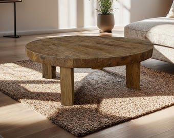May include: A round, rustic wooden coffee table with a natural finish. The table sits on four sturdy, square legs and is placed on a woven, beige rug. The room has a neutral color palette with natural light.