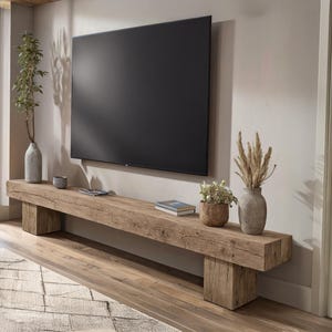 May include: A rustic, wooden TV stand with a large, black screen mounted above it. The stand features a natural wood finish and is decorated with vases, plants, and books. The room has a neutral color palette with a rug and hardwood flooring.