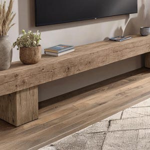 May include: A long, rectangular wooden console table featuring a natural, weathered grain finish. The piece is supported by two thick, block-style legs and holds a tall gray vase with dried grass, a small potted plant, and a stack of books.