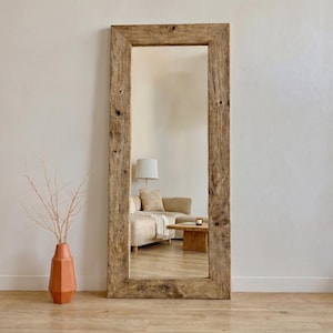 Puede incluir: Un espejo de cuerpo entero rectangular grande con un marco de madera rústico y desgastado. El espejo refleja un salón con un sofá beige, una mesa de centro de madera y una lámpara. Un jarrón de terracota con ramas secas está a la izquierda.