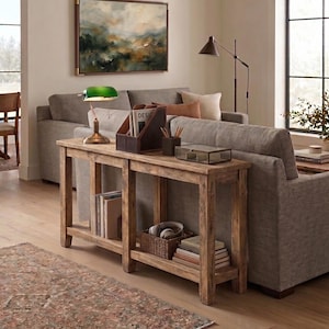 May include: A rustic wooden console table with two shelves, holding books, a woven basket with headphones, and decorative items. A green banker's lamp and a brown leather magazine holder sit on top. A gray sofa and abstract landscape painting are in the background.