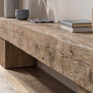 May include: A long, rectangular bench crafted from reclaimed wood with a prominent, weathered grain and visible natural cracks. The bench features a thick, solid top supported by sturdy, block-style legs, showcasing a raw, textured finish.