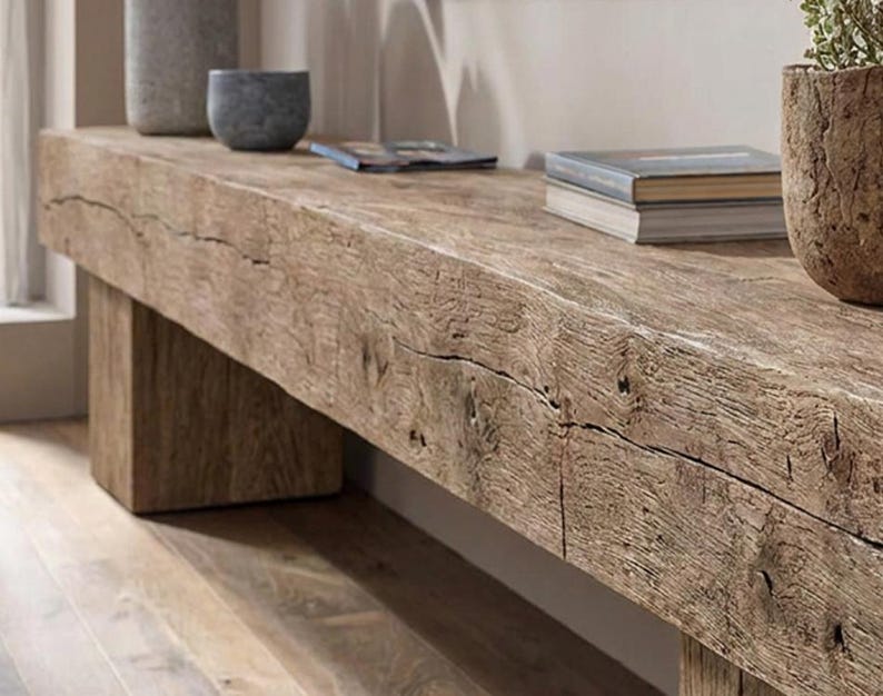 May include: A long, rectangular bench crafted from reclaimed wood with a prominent, weathered grain and visible natural cracks. The bench features a thick, solid top supported by sturdy, block-style legs, showcasing a raw, textured finish.