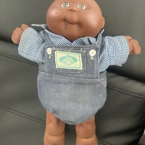 Puede incluir: Muñeca vintage Cabbage Patch Kids con piel morena, vestida con una camisa a cuadros azul y blanco y un peto vaquero. La muñeca tiene ojos marrones y una expresión sonriente. El logo de Cabbage Patch Kids es visible.