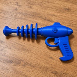 Peut inclure: Un pistolet jouet bleu vif doté d'un long canon avec quatre ailettes circulaires, d'un corps principal arrondi avec un petit bouton circulaire sur le côté, d'un pontet et d'une poignée texturée avec des rainures horizontales.