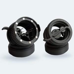 Könnte beinhalten: Zwei schwarze Espresso-Kaffeeverteilungswerkzeuge mit kreisförmigem Design. Jedes Gerät verfügt über einen strukturierten Außenring, interne Zahnradmechanismen und eine Reihe dünner Metallnadeln zum Bearbeiten des Kaffeemehls.
