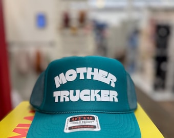 Könnte beinhalten: Eine türkisblaue Trucker-Kappe mit weißem Netzrücken. Die Vorderseite der Kappe trägt den Text "MOTHER TRUCKER" in Weiß, umrandet von Schwarz. Ein kleines weißes Etikett mit schwarzem Text ist an der Vorderseite der Kappe befestigt.