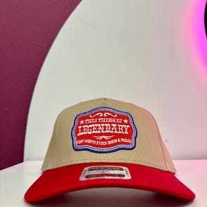 May include: A tan baseball cap with a red brim. The cap features a patch with the words "THIS THING IS LEGENDARY" and "FORT WORTH STOCK SHOW & RODEO" in a red and blue design.