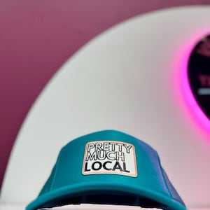 May include: A teal trucker hat with a mesh back and a curved brim. The hat features a rectangular patch with the text "PRETTY MUCH LOCAL" in a bold, sans-serif font. The patch has a white background with a peach-colored border.