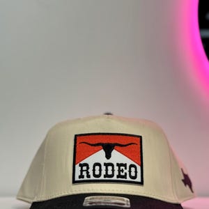 May include: A beige and black baseball cap with a rectangular patch showing a bull's head and the word "RODEO". The cap has a black peak and a star on the side.