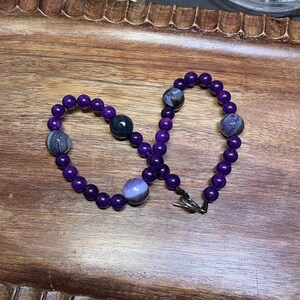 May include: A purple beaded necklace with a silver clasp. The beads are made of amethyst and agate, and they have a variety of patterns and colors.
