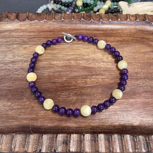 May include: A purple and yellow beaded necklace with a silver toggle clasp. The necklace is made of round beads and is approximately 18 inches long.