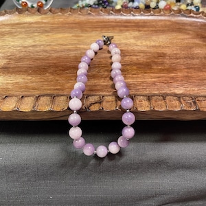 May include: A purple and white beaded necklace with a silver clasp. The necklace is on a wooden surface.