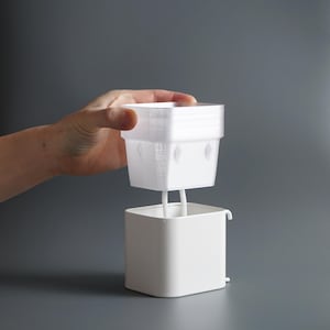 May include: A white plastic self-watering planter with a removable top. The top has a square shape and a textured surface. The bottom of the planter is square with a rounded edge.