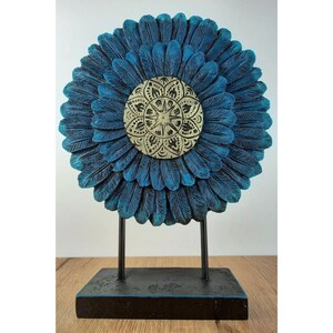 Könnte beinhalten: Eine blaue Holzblumen-Skulptur mit einem goldenen Mandala-Design in der Mitte. Die Skulptur steht auf einem schwarzen Holzsockel.