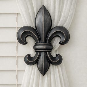 May include: A dark gray fleur-de-lis curtain tieback. The decorative metal accent features a stylized lily design with three curved petals and a central vertical element. The tieback is holding a white curtain in place.
