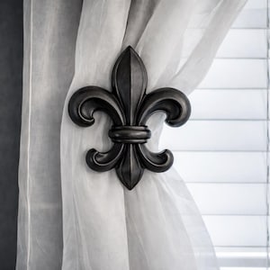 May include: A dark gray fleur-de-lis curtain tieback. The decorative metal accent is in the shape of a stylized lily flower, holding sheer white curtains. The background includes a window with white blinds.
