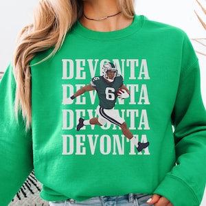 May include: A bright green sweatshirt featuring the name "DEVONTA" stacked vertically with a football player graphic. The player is wearing a green and white uniform and running with a football. The sweatshirt is a crew neck style.