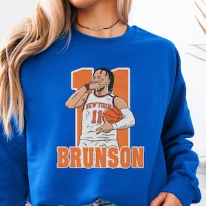 May include: A royal blue sweatshirt with a graphic of a basketball player wearing a white and orange jersey with the text "NEW YORK" and "11" on the front. The text "BRUNSON" is printed in orange below the graphic.