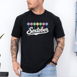 May include: A black short-sleeved t-shirt featuring a graphic of seven colorful swirled lollipops arranged in a row above the white text "Soxtober" written in a cursive script font with a decorative underline.