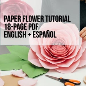 May include: A large, light pink paper flower with layered petals and green paper leaves. The image includes text that reads: PAPER FLOWER TUTORIAL 18-PAGE PDF ENGLISH + ESPAÑOL. A pair of black and orange scissors rests on the surface nearby.