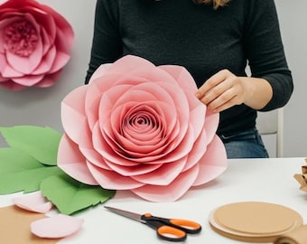 Peut inclure: Une grande rose en papier rose clair avec des pétales détaillés. Des feuilles de papier vertes et des pétales roses sont disposées sur une surface blanche. Des ciseaux avec des poignées orange et du matériel de bricolage sont également visibles.