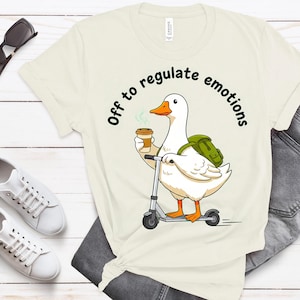 Puede incluir: Camiseta color crema con un pato de dibujos animados en un scooter, sosteniendo una taza de café. El pato lleva una mochila y el texto "Off to regulate emotions" está arriba. Gafas de sol y zapatillas blancas también son visibles.