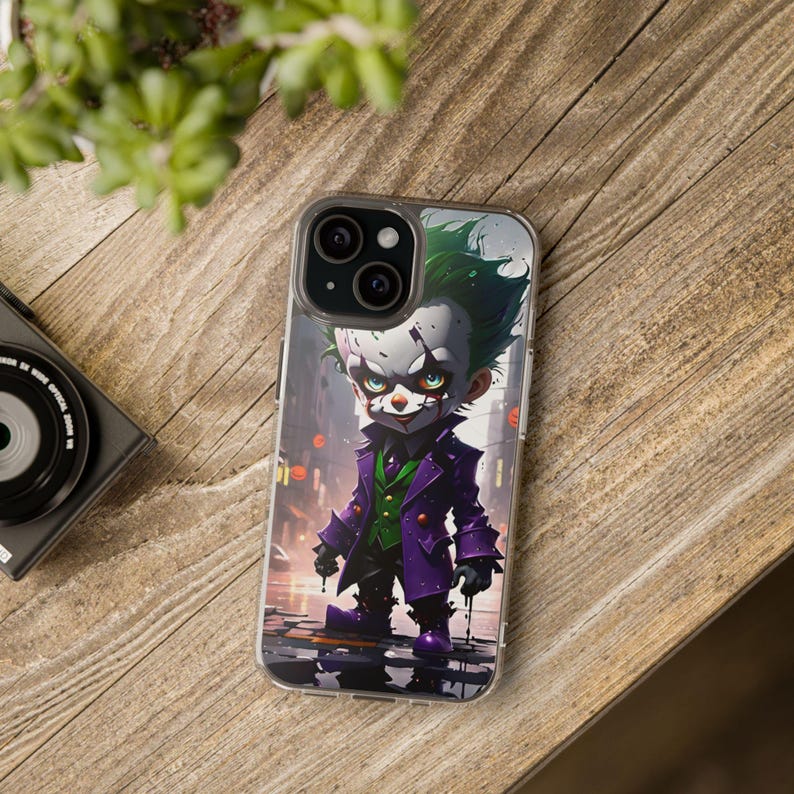 May include: A clear phone case featuring a colourful illustration of the Joker from Batman. The Joker is depicted as a small, cartoonish character wearing a purple and green outfit, with a mischievous grin and white makeup.