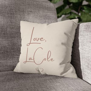 May include: A square throw cushion in a light beige colour featuring the text Love, LaCole in a dusty rose cursive font. Below the text is a small, simple outline of a heart shape. The cushion has a smooth fabric texture and a clean, square shape.