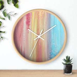 May include: Circular wall clock with a light wood surround. The clock face displays a colourful abstract design with vertical stripes in pink, yellow, and blue. White clock hands show the time. A decorative item for any room.