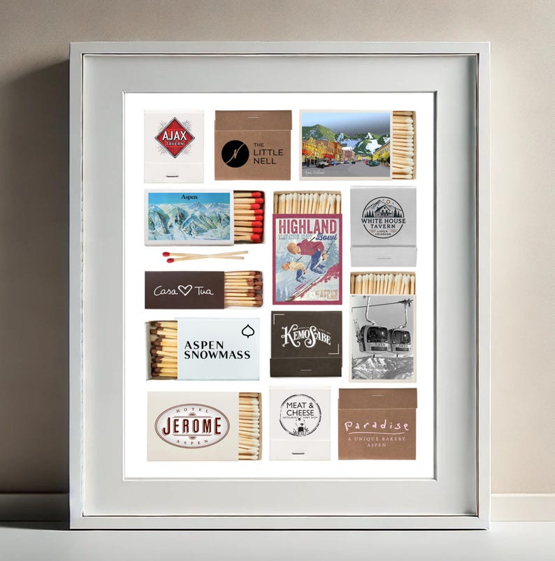 May include: A framed collage of matchbox labels featuring various ski resorts and businesses in Aspen, Colorado. The labels are arranged in a grid pattern and include names like "Ajax", "The Little Nell", "Highland Bowl", "White House Tavern", "Casa Tua", "Aspen Snowmass", "Kemo Sabe", "Meat & Cheese", "Jerome", and "Paradise".