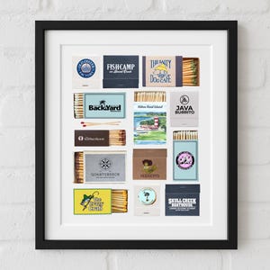 Puede incluir: Una impresión enmarcada con varios diseños de cajas de cerillas. Cada caja de cerillas tiene un diseño y un texto únicos, incluyendo "Old Oyster Factory", "Fishcamp", "The Salty Dog Cafe", "Back Yard", "Java Burrito" y "Skull Creek Boathouse".