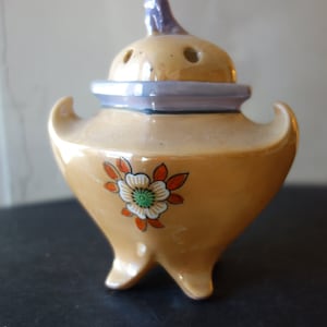 Puede incluir: Un pequeño quemador de incienso de cerámica con un diseño floral. El quemador es de color marrón claro con un borde morado y una flor con detalles en naranja y verde. Tiene tres patas y una pequeña abertura en la parte superior.