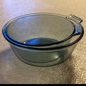 May include: A set of two clear glass mixing bowls with a blue tint. The bowls are nested inside each other. The larger bowl has a handle on the side.