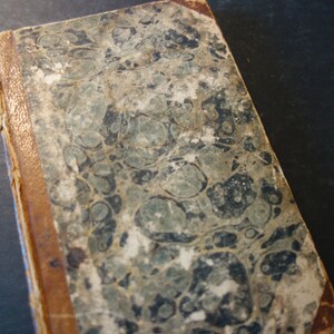 May include: A vintage book with a marbled cover in shades of brown, black, and white. The book has a worn leather spine with gold accents.