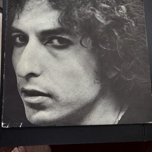 May include: Black and white album cover featuring a close-up portrait of a man with curly hair. The album title "Hard Rain" and the artist's name "Bob Dylan" are printed in white text.
