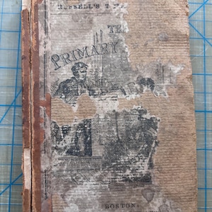 以下が含まれることがあります： 古書。 擦り切れたベージュ色の表紙と目に見える損傷があります。「PRIMARY」のタイトルが大きく表示され、人々のイラストが描かれています。 背表紙は茶色で、表紙には経年劣化と摩耗の兆候が見られます。