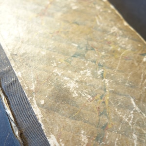 Pode incluir: Close-up de um livro antigo com uma capa desgastada e texturizada. A capa tem um padrão marmorizado em tons de bege, castanho e toques de azul e amarelo. A lombada é encadernada com um tecido canelado cinza escuro.