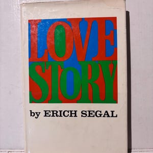 以下が含まれることがあります： エリック・シーガルの「LOVE STORY」というタイトルのヴィンテージペーパーバック。表紙には、赤、緑、青の大きな太字のカラフルな文字でタイトルが書かれています。本は白い背景に著者の名前が黒で書かれています。