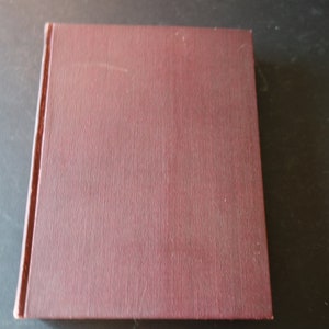 Puede incluir: Un libro rectangular cerrado con una cubierta burdeos. La cubierta tiene un aspecto texturizado y un ligero brillo. El lomo es visible en el lado izquierdo, y el libro está sobre una superficie oscura.